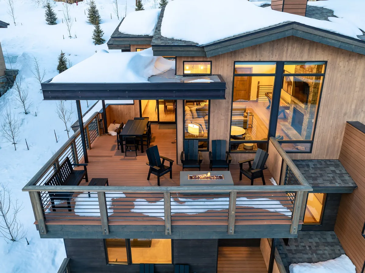 The image offers a view of a cozy outdoor setting with a fire pit, outdoor seating, and a dining table, all positioned in front of large windows. Snow blankets the surroundings, highlighting the villa as a welcoming base for winter fun and group experiences.