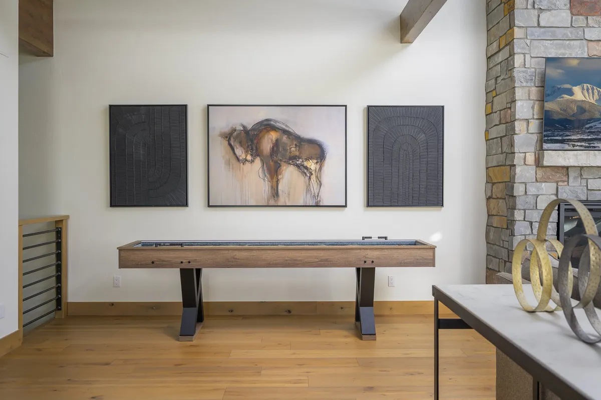 A lively corner of the gathering area showcasing a shuffleboard table, tasteful buffalo-themed artwork, geometric wall art, and a stone fireplace, all contributing to a playful yet elegant setting. The space feels ready for both friendly competition and relaxed socializing.