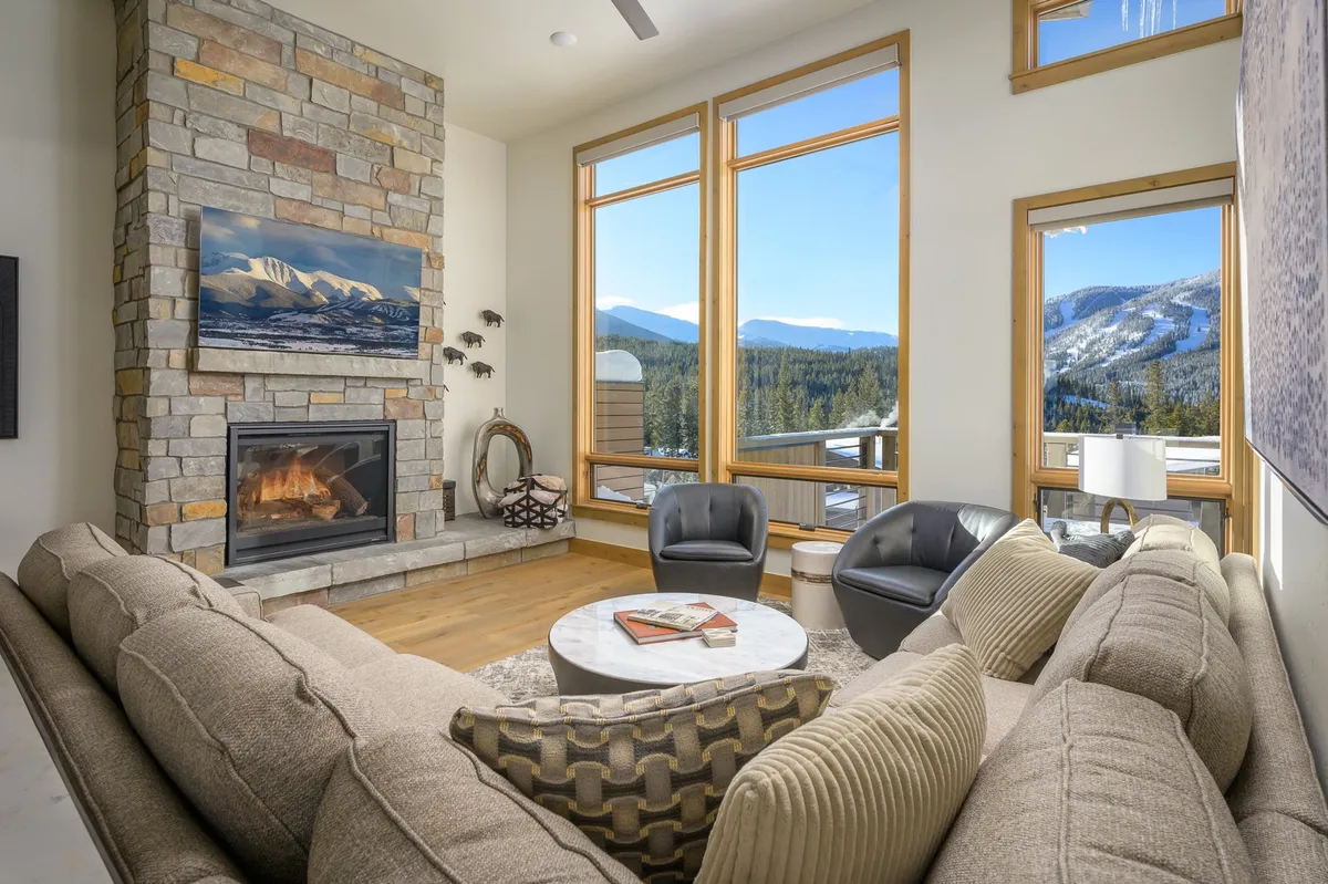 A spacious living room featuring a striking stone fireplace, plush comfortable seating arranged around a central coffee table, and expansive windows that perfectly frame a dramatic mountain vista. Sunlight pours through the windows, illuminating the warm textures and inviting ambiance of the space.