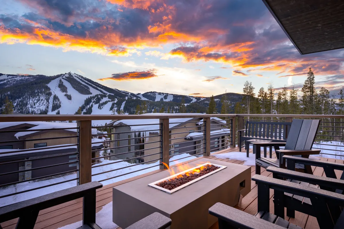 An expansive exterior deck featuring cozy seating around a fire pit, overlooking a breathtaking mountain view. The sunset sky bathes the space in warm light, inviting relaxation or gatherings under the stars.