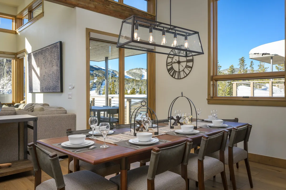 A bright, inviting dining area furnished with a custom wooden table, statement chandelier, and large windows that open up to sweeping mountain views. The space is arranged for group gatherings, celebrations, or casual meals, with sunlight highlighting the warmth of the natural materials.