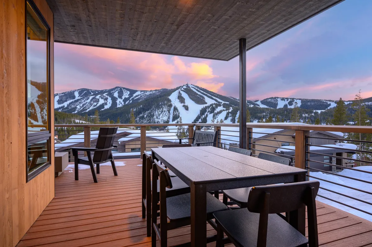 An outdoor dining table is set on a deck with a clear mountain view, framed by deck railings. The setting suggests al fresco meals, social gatherings, and uninterrupted enjoyment of the surrounding scenery.