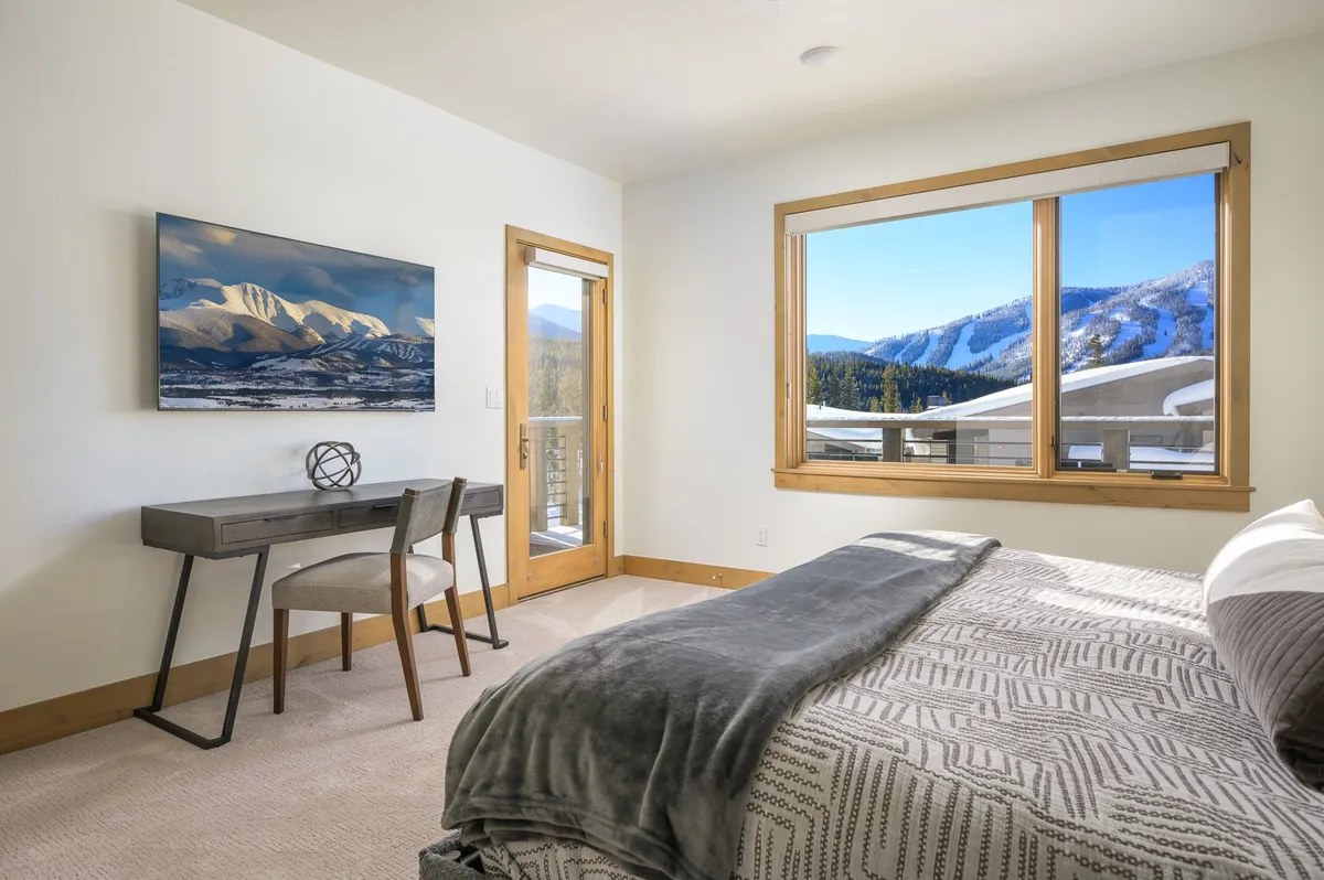 A serene guest bedroom featuring a large window with mountain views, a comfortable bed, and a stylish desk and chairs, blending relaxation with the option for quiet productivity.