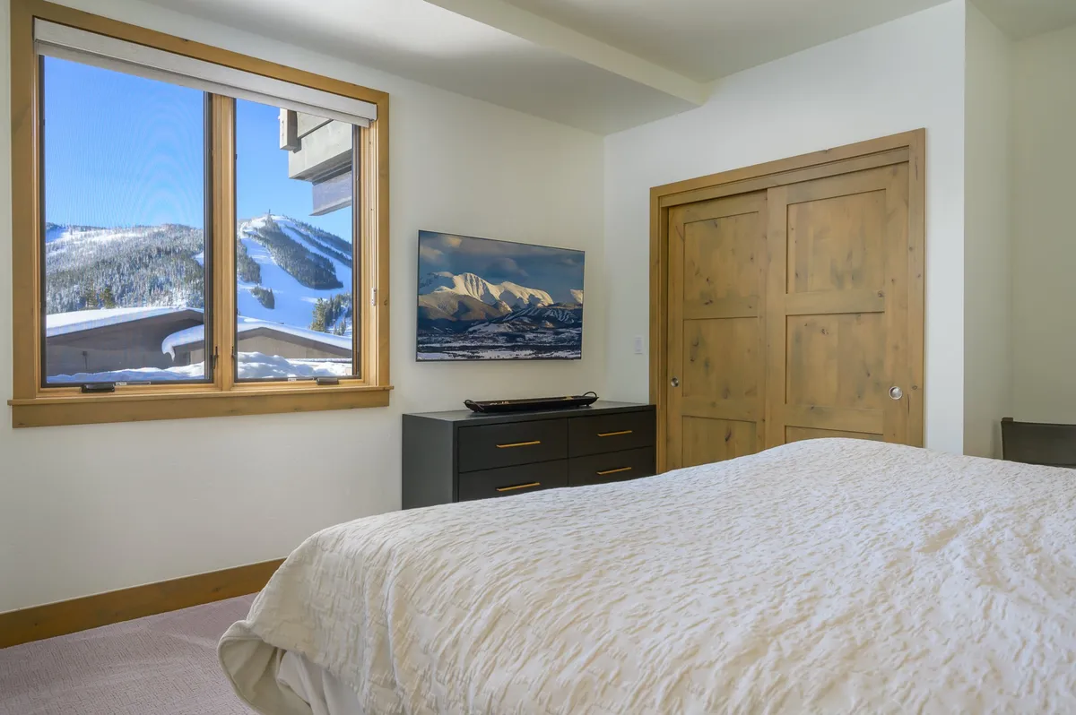 A tranquil bedroom with a large window overlooking the mountains, inviting bed with textured bedding, and a TV displaying a mountain scene, evoking restfulness and warmth.