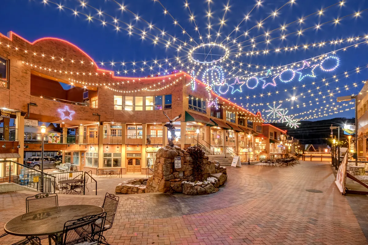 A lively outdoor shopping and dining area illuminated by string lights and festive decorations. People gather at outdoor seating, surrounded by unique shops and a stone fountain, capturing the vibrancy of the community across seasons.